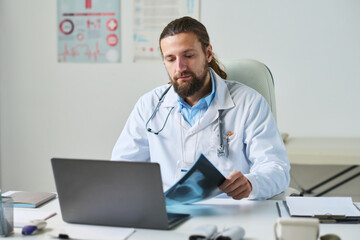 Confident male general practitioner in lab coat looking at x-ray while consulting online patient by workplace in medical office in clinics