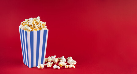 Blue white striped carton bucket with tasty cheese popcorn, isolated on red background. Box with scattering of popcorn grains. Fast food, movies, cinema and entertainment concept.