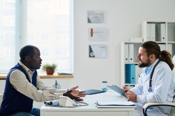 Fototapeta na wymiar Mature African American man describing his symptoms to young doctor listening to him and making notes during medical consultation