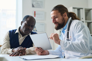 Fototapeta premium Serious mature patient looking at tablet screen and listening to clinician explaining results of medical examination during consultation
