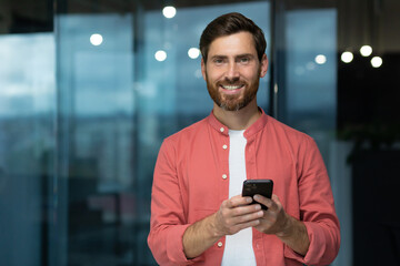 Portrait of a young man in a red shirt standing in the office, holding a phone in his hands, looking at the camera with a smile.