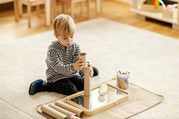 A little boy is sitting on the floor and building a tower while playing with montessori toy in kindergarten.