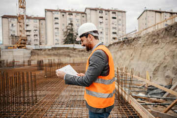 A happy engineer is holding plans and documents and checking on it while standing at construction...
