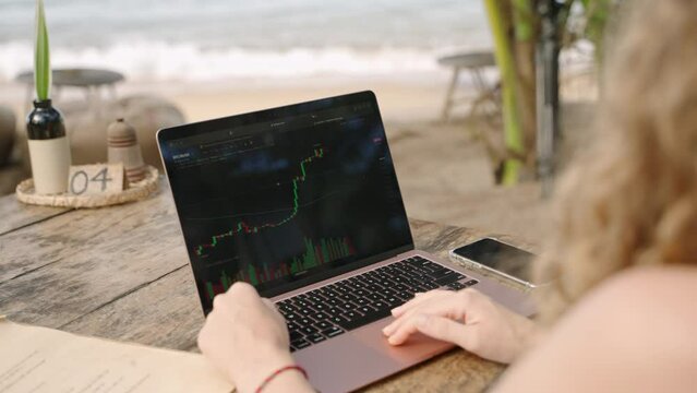 Female cryptocurrency trader at laptop checking charts online working remotely at outdoor tropical seaside cafe. Woman crypto broker analyses graphic of stock exchange rates and bets by ocean close-up