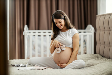 A pregnant woman is sitting on a bed with baby shoes in her hands and cuddling tummy.