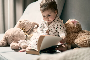 Fototapeta na wymiar A little baby sitting on the sofa reading book. Teddy bear and rabbit toys are nearby