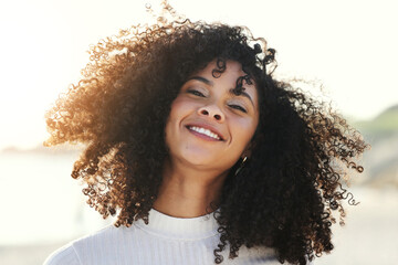 Black woman, afro and wind in hair outdoor with a smile and portrait at beach for vacation or freedom. Face of happy young model person in nature for peace, travel and time to relax on sunset holiday