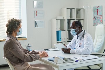 Experienced clinician with tablet and mature female patient wearing protective masks during communication at medical consultation