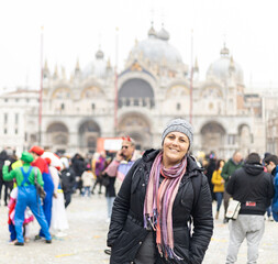 Obraz premium Woman in winter clothes and hands in pockets smiling at San Marco Square, Venice. Crowd and basilica on the background
