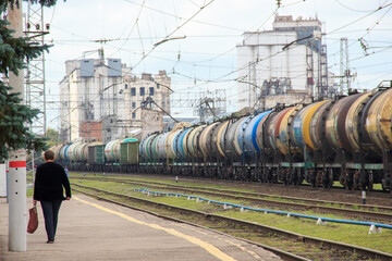 a female figure walking down a sidewalk next to a train and railways with grass.huge number of wagons with oil products, wires in the sky, an old factory