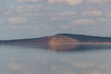 lake with clouds in the sky, red hill with forests on background. hill symmetric reflection in the water