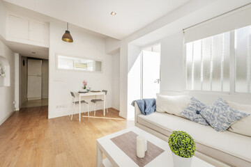 Living room of a loft-style rental house with a white three-seater sofa, a matching coffee table, a barred window and an exit door to a terrace
