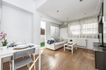 Living room of a house furnished with a three-seater sofa in white fabric, doors leading to a terrace, windows with bars and a white square dining table