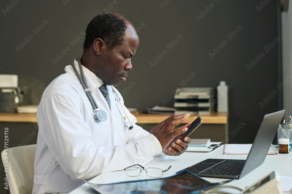 Aufkleber side view of serious mature clinician scrolling in smartphone while sitting in front of laptop by wo
