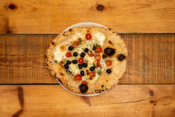 A stone-baked Italian pizza on a wooden table with black olives, cherry tomatoes, basil and oregano