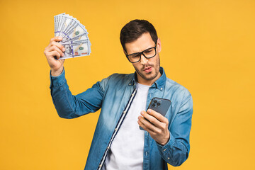 Happy winner! Excited man in casual holding lots of money in dollar currencys and using phone in hands isolated over yellow background. - 577131504