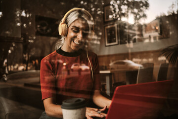 Woman smiling while using headphones and laptop in a coffee shop.