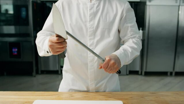 A chef is sharpening a knife at a wooden table in a professional kitchen