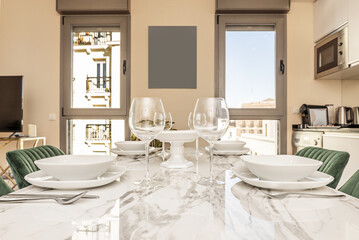 A white marble dining table with crystal stemware and white crockery in a room with an open plan kitchen and twin windows on one wall