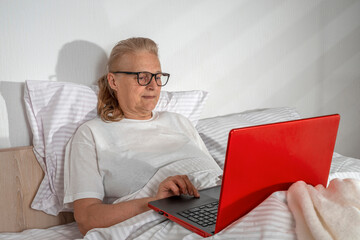 Senior woman using laptop lies in bed in bedroom. Lifestyle. Online activity in old age, bright mind, technologies