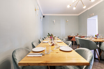 Long light wood dining table with food service set with green velvet upholstered stools in a bar dining room with multi-bulb lamps, light green walls and gray tiled floors