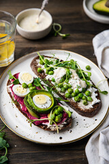 Two smorrebrods (open sandwiches) with different toppings: green peas - cottage cheese and quail eggs -beetroot paste- avokado- cream cheese on rye bread and glass of lemonade on wooden table. 