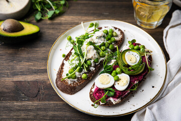 Two homemade smorrebrods (open sandwiches) with different toppings: green peas-cottage cheese and quail eggs-beetroot paste- avokado- cream cheese on rye bread and glass of lemonade on wooden table. 
