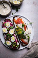 Two smorrebrods (open sandwiches with different toppings: avocado-salmon-cream cheese and quail eggs -beetroot - cream cheese) and mustard sauce on bright surface.