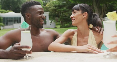 Happy diverse couple wearing swimming suits and drinking drinks at swimming pool in garden