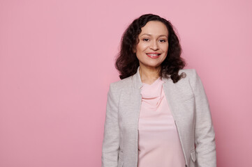Fototapeta premium Portrait of a confident dark-haired middle-aged multi-ethnic woman in elegant light gray jacket, smiling a toothy smile, looking at camera, isolated on pink background with copy space. People concept