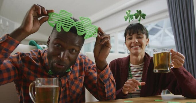 Portrait of happy diverse couple with beer wearing clover shape items and having video call