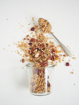 Top view of delicious sweet granola with assorted nuts and dried fruits spilled from glass on white table near metal spoon