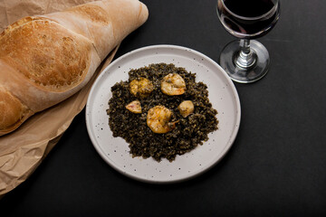 Black risotto with squid ink and shrimp, served on a white plate accompanied by a glass of wine and freshly baked bread. Italian cuisine. Black rice.