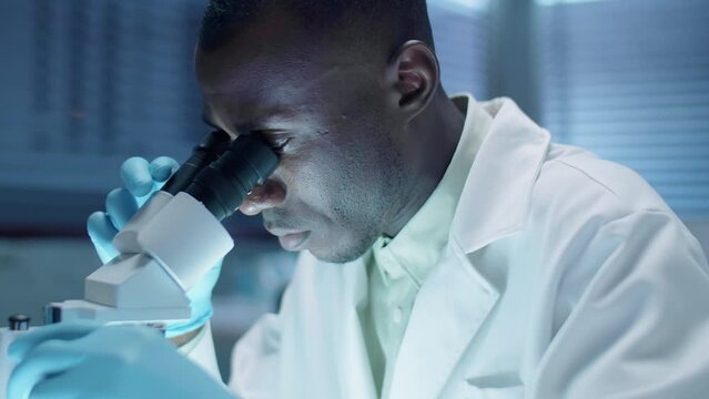 Medium close up shot of black scientist in sterile gloves and white coat using microscope and making notes while doing research in laboratory
