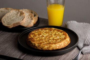 Traditional Spanish omelette with egg and onion served on a black plate, accompanied by a glass of orange juice and bread.