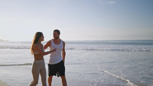 Young biracial happy couple holding hands and walking on the beach together enjoying summer sideview. Cheerful boyfriend and girlfriend relaxing and talking at seaside hugging and kissing at sunrise.