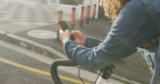 Happy biracial woman in city, sitting on bike using smartphone