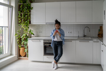 Full length of Asian woman housewife holding mobile phone using app to pay utility bills. Millennial girl standing in kitchen at home looking at smartphone screen searching cooking recipes