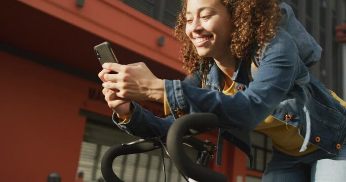 Happy biracial woman in city, sitting on bike using smartphone