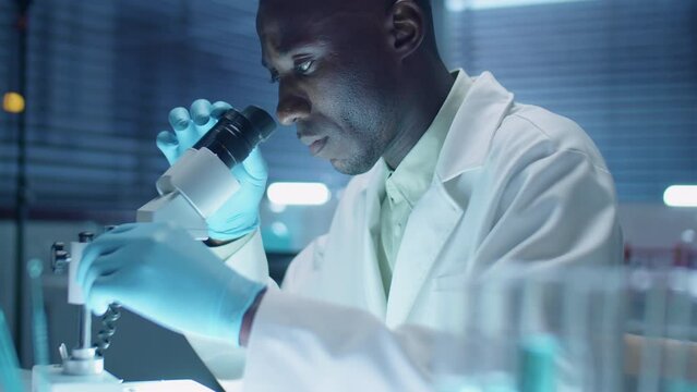 Black scientist in sterile gloves and lab coat looking through microscope while doing research in laboratory