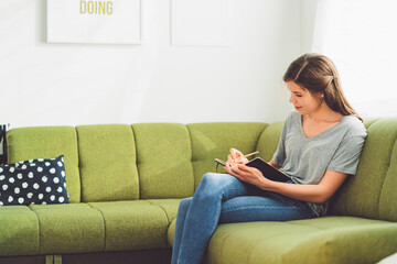 Relaxed young woman writing in her notebook, sitting on the green couch
