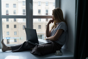 A young woman sits on a windowsill, works at a laptop, looks out the window.