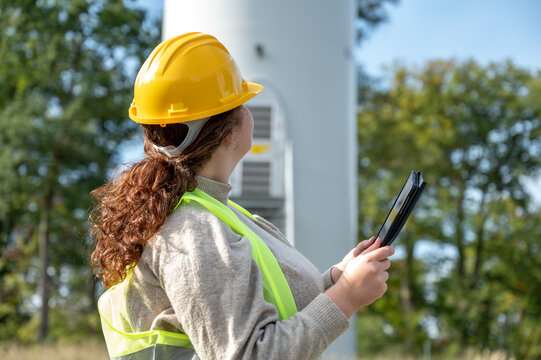 Female engineer with brown curly hair, yellow helmet and warning vest using a digital tablet to analyse the statistics of a modern renewable energy wind turbine, rear view