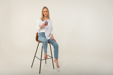 Fototapeta na wymiar Full length side profile photo of young attractive girl in jeans and white shirt looking, browsing mobile phone while sitting on stool isolated on white background