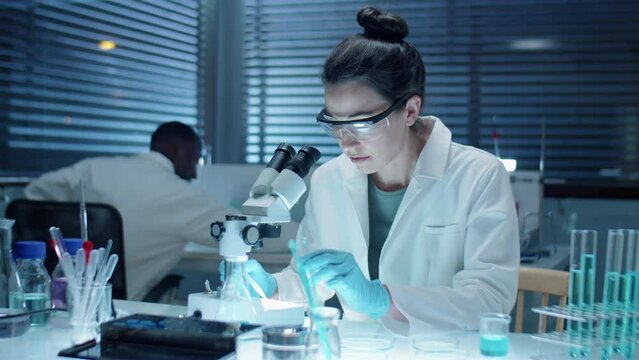 Medium shot of female scientist in sterile gloves, protective glasses and white coat examining chemicals under microscope while working late in laboratory