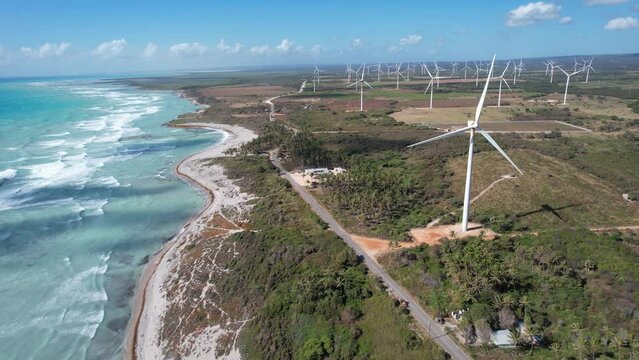 Beautiful video of a wind farm is seen towering over a stunning beach. The wind turbines, with their sleek white blades, stand tall against the clear blue sky, while the ocean waves lap gently a