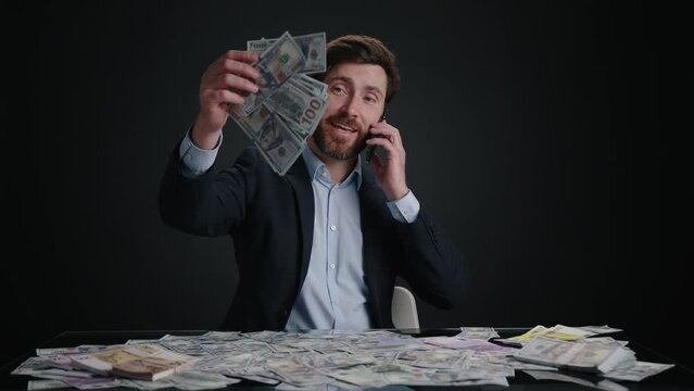 A man in a suit dropping money while talking with someone on the phone. He looks happy. The man is in black room. The m,oney is in a big pile. High quality 4k footage 