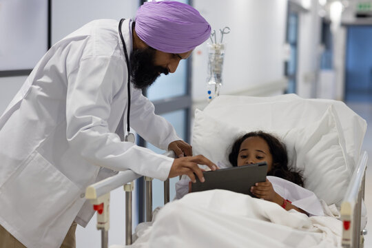 Sikh male doctor in turban with girl using tablet and lying on hospital bed - Powered by Adobe