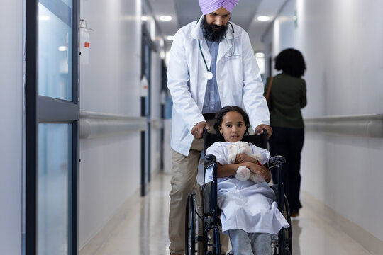 Portrait of sikh male doctor in turban walking with girl in wheelchair in hospital