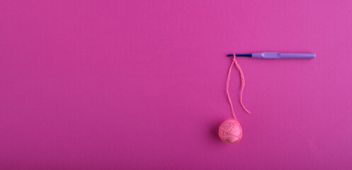 A banner with a blue shiny hook with a light purple plastic handle, on which a chain of loops from a ball of soft orange threads is typed, on a bright pink textured background. Knitting, needlework.